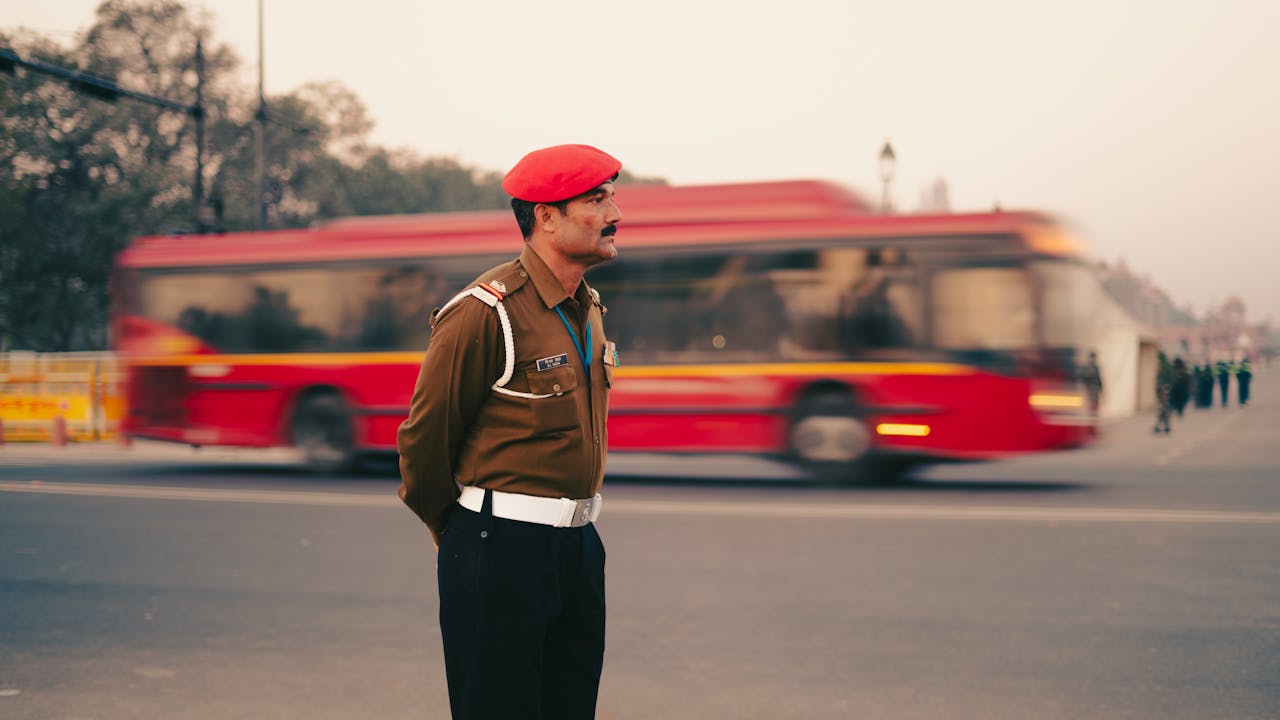 A traffic officer in a red beret stands in New Delhi, with a blurred red bus in the background.